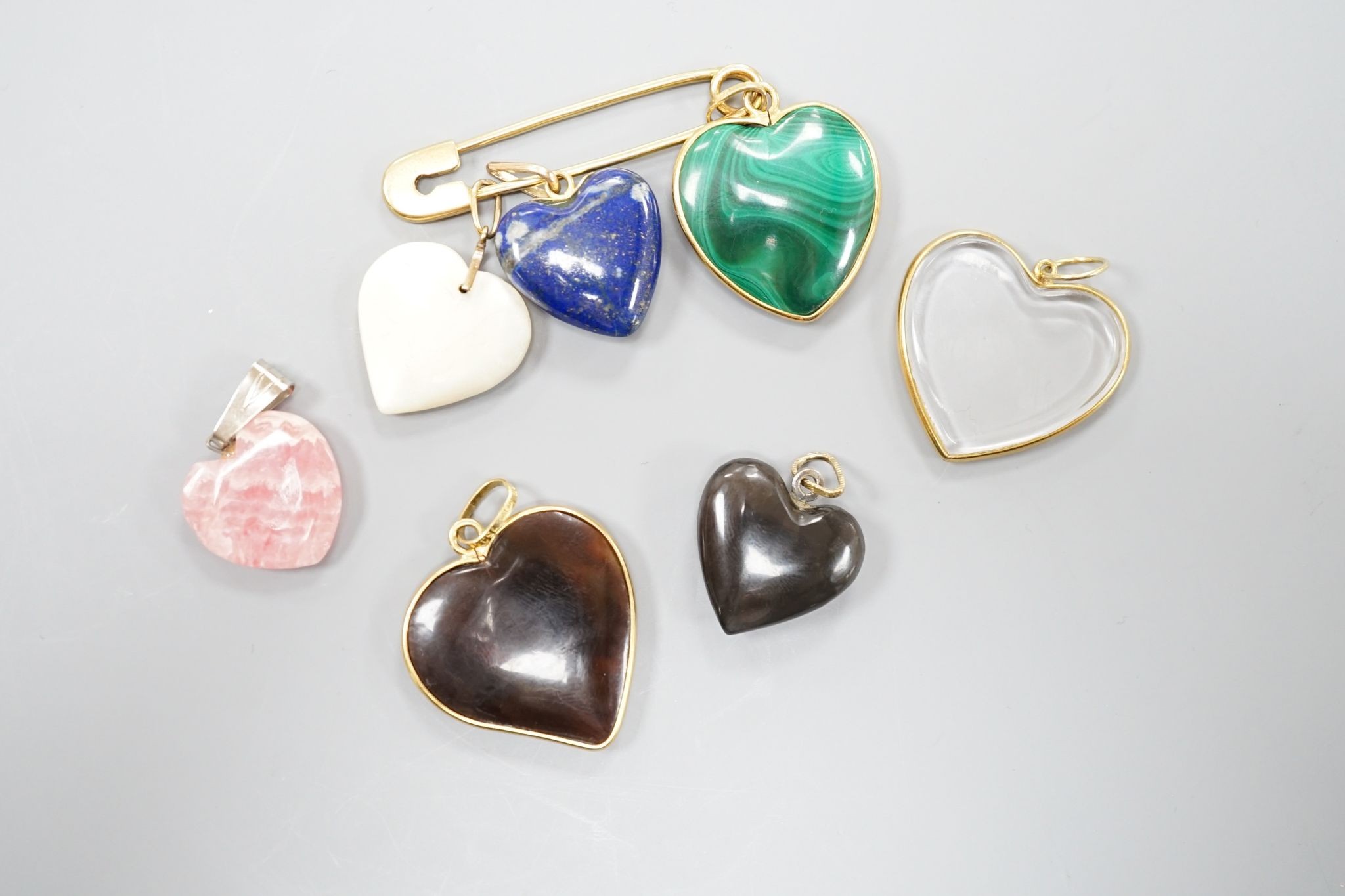 An 18ct safety pin, 47mm, hung with seven assorted heart shaped hardstone pendants or charms, two with Italian 750 yellow metal mounts.
